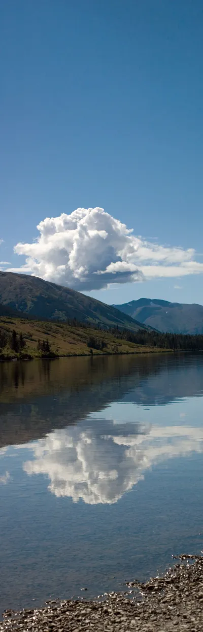A vertical panoramic view of a mountain reflecting in the calm, clear water of a lake in Cooper Landing, with a large white cloud in the blue sky.