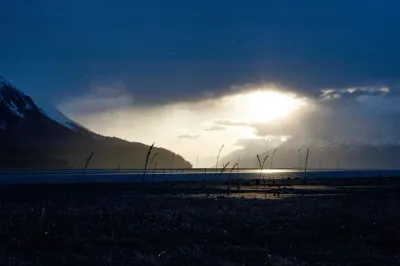 A moody sunset view of Turnagain Arm at low tide, with the sun breaking through dramatic clouds and reflecting on the wet mudflats.