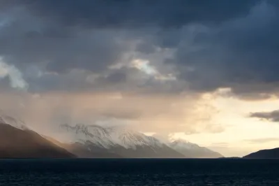 A moody evening shot of snow-dusted mountains across Turnagain Arm, with the last rays of sun hitting the slopes under a stormy sky.