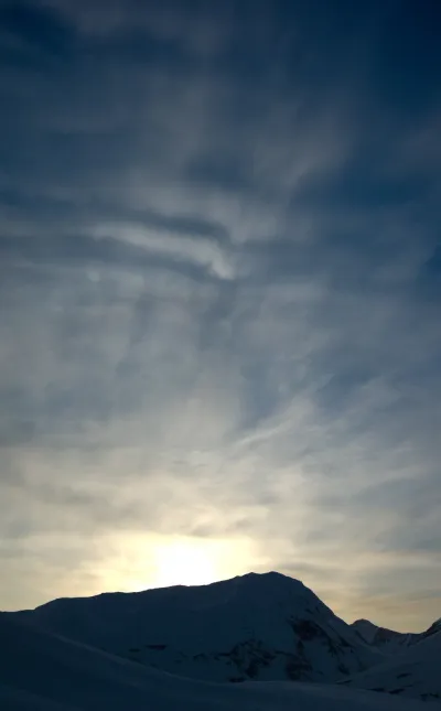 A majestic view of a silhouetted mountain peak at sunset, with a vibrant golden glow on the horizon and cirrus clouds in the sky.