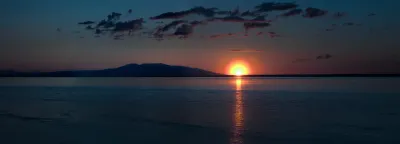 A panoramic sunset over the ocean, with the sun on the horizon reflecting on the water and a mountain silhouette in the distance.
