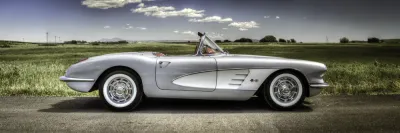 A classic silver 1960 Corvette convertible with white sidewall tires parked on the side of a rural road under a sunny sky.