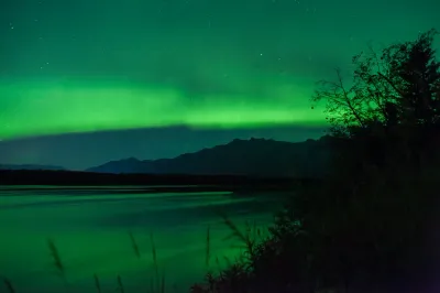 A vivid green aurora borealis streaks across the night sky above a dark mountain range and a reflecting body of water. The aurora is reflected in the water, creating a symmetrical glow.
