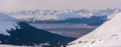 A snow-filled valley viewed between two steep mountain slopes in Alaska, with a river or mudflat visible in the distance.