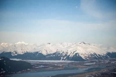 A wide panoramic view of snow-covered mountains in Alaska, with a large river or mudflat stretching through the valley under a clear blue sky.