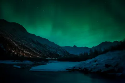 A wide field of green aurora light hangs above a rugged, snow-dusted mountain valley. A partially frozen river or lake reflects the green glow in the foreground.