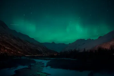 A view of the green aurora borealis appearing as soft green clouds scattered across a starry sky over a snow-dusted mountain valley.