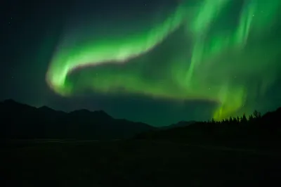 A dramatic green aurora arc sweeps across the sky above a dark silhouetted mountain range, showing vibrant vertical light structures.