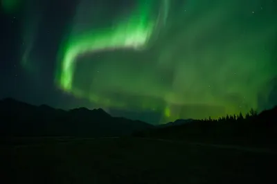 A vibrant green aurora arc over silhouetted mountains, with a slightly different light pattern than nearby shots.