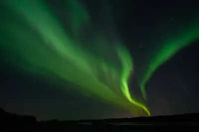 Vibrant green aurora curtains with intense light towards the horizon, arching over a dark mountain silhouette.
