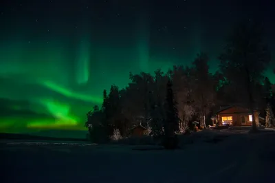 A cozy, illuminated wooden cabin sits amidst a snowy forest under a sky filled with green aurora light. The warm light from the cabin windows contrasts with the cool green and blue of the night.