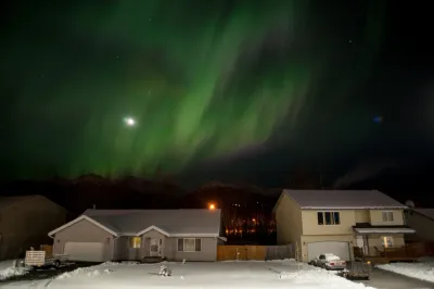 Vibrant green aurora light dances over a residential neighborhood at night, with two-story houses in the foreground and mountains in the distance.