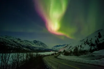 A bright swirl of green and pink aurora light dominates the sky above a winding road through a snowy mountain pass.