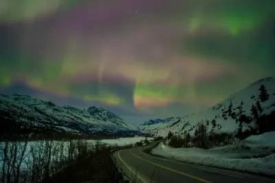A broad view of green and pink aurora curtains over a snowy mountain road, with soft and blended colors.
