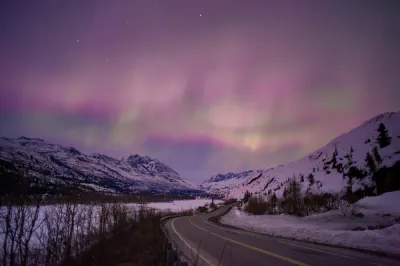 The aurora appears with distinct pink and purple hues, forming ethereal curtains over a snowy landscape and winding road.