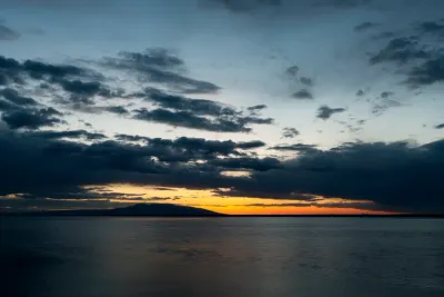 A sunset view over the water, with silhouetted mountains on the horizon and a dramatic, cloudy sky.
