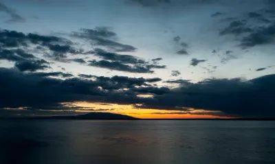A vibrant sunset over a calm body of water, with silhouetted mountains in the background and clouds catching the golden light.