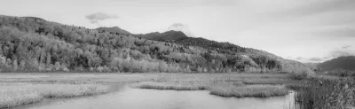A black and white panoramic view of a vast wetland with grassy islands and a calm water surface reflecting a forested hillside and distant mountains.