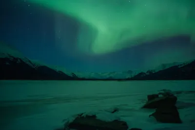 A soft, diffused green aurora glow hangs low over a frozen landscape with dark mountain silhouettes under a clear, starry sky.