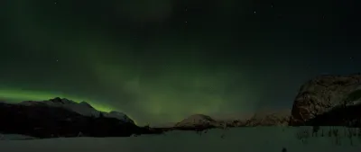 A wide panorama of multiple snowy peaks under a dark sky with a faint, reaching green aurora light.