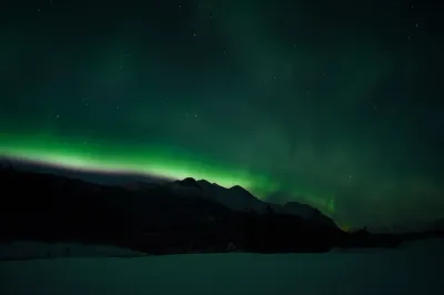 A faint green aurora light reaches across a dark sky above a silhouetted mountain range and a vast, snowy foreground.