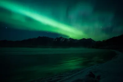 A bright, concentrated green aurora beam stretches across the sky above a jagged mountain range, reflecting in a partially frozen river below.