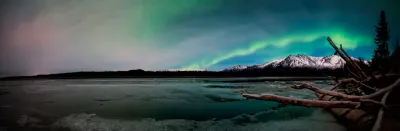 A panoramic view of a wide band of green aurora arcing over a large frozen lake and distant snow-capped mountains, with a fallen tree trunk in the foreground.