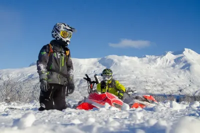 Two snowmobilers in full gear standing next to their machine in a deep, snowy field.