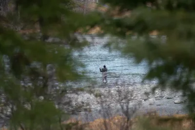 A lone fisherman wading in a turquoise river, framed by the blurry branches of trees in the foreground.