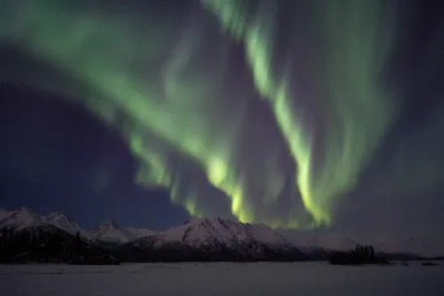 A broad green aurora arc hangs over a snowy mountain range, with vibrant light vertical structures.