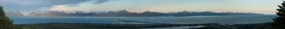 A wide panoramic view of Kachemak Bay from Homer, Alaska, featuring the Homer Spit and the snow-capped Kenai Mountains under a clear blue sky.