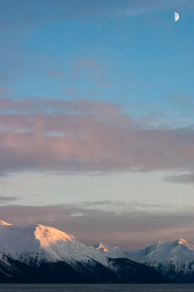 A beautiful sunset view from an elevated position in Alaska, with a crescent moon in the sky and warm light hitting the snow-covered mountains.