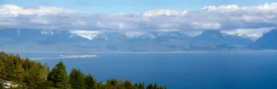 A wide panoramic view of Kachemak Bay from Homer, Alaska, featuring the Homer Spit and the snow-capped Kenai Mountains under a clear blue sky.