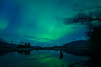 A scenic view of a calm lake reflecting the starry night sky and a soft green aurora glow, with snow-capped mountains in the background.