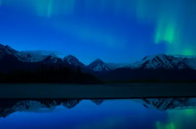 A calm lake reflection with more pronounced green and blue aurora light over the distant snowy peaks.