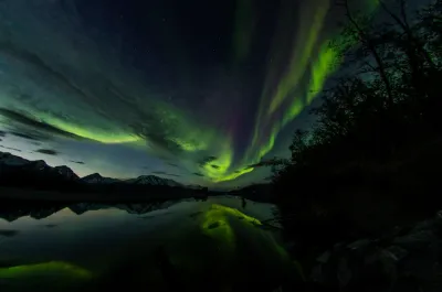 A vibrant green and purple aurora curtain swirls beautifully over a reflecting lake and dark mountain silhouettes.