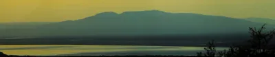 A wide panoramic view of a silhouetted Alaskan mountain range under a pale yellow sky at sunset.