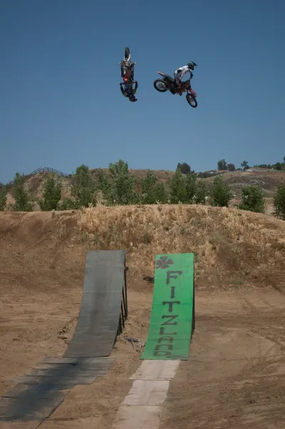 Two freestyle motocross riders performing high-flying tricks simultaneously in the air above ramps labeled 'FITZLAND'.
