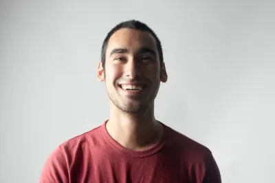 A close-up portrait of a smiling young man in a maroon t-shirt against a plain grey background.
