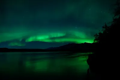 A wide field of green aurora light hanging above a dark mountain range and reflecting in a body of water.
