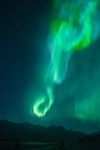 A tall, swirling column of green aurora light hangs high above a dark mountain range.