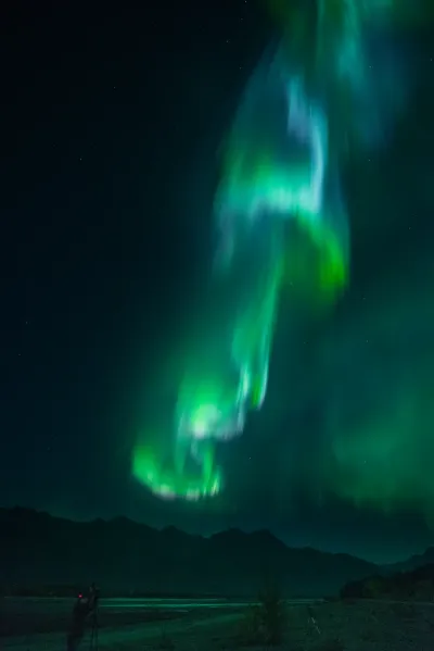 An intense column of green and purple aurora light stretching over a dark mountain skyline.