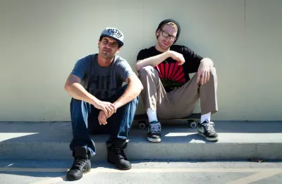 A photo of two friends sitting on a curb, one on a skateboard, with a casual, cool vibe.