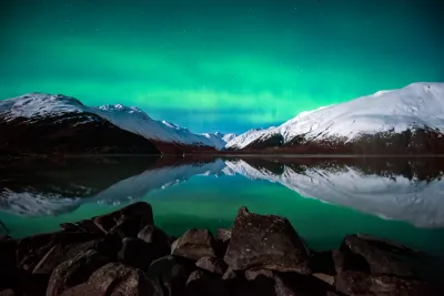 A green aurora band reflecting in a perfectly calm, icy lake surrounded by snow-covered mountains, with dark boulders in the foreground.