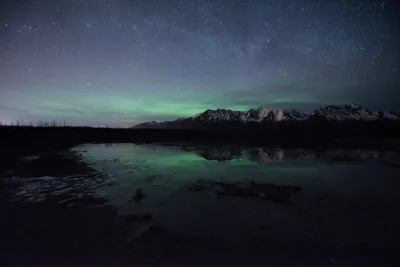 A wide field of soft green aurora under a bright starry sky, with the Milky Way faintly visible above a dark mountain range.