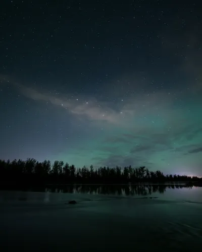 The starry sky and a soft green aurora glow over a calm lake and forest silhouette.