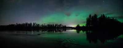 A panoramic shot of a wide green aurora band over a calm river and dark forests under a brilliant starry sky.