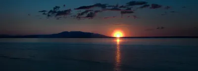 A vibrant sunset over a calm body of water, with silhouetted mountains in the distance and a fiery orange-red glow on the horizon.