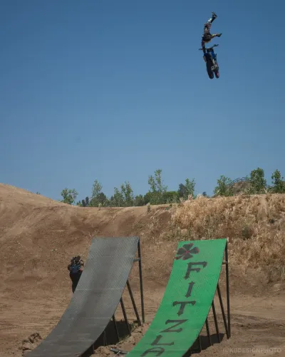 A freestyle motocross rider performing a massive backflip trick high in the air above a ramp labeled 'FITZLAND'.