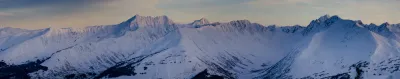 A wide-angle panoramic view of a snow-filled valley and jagged, snow-covered mountains in Turnagain Pass, Alaska.